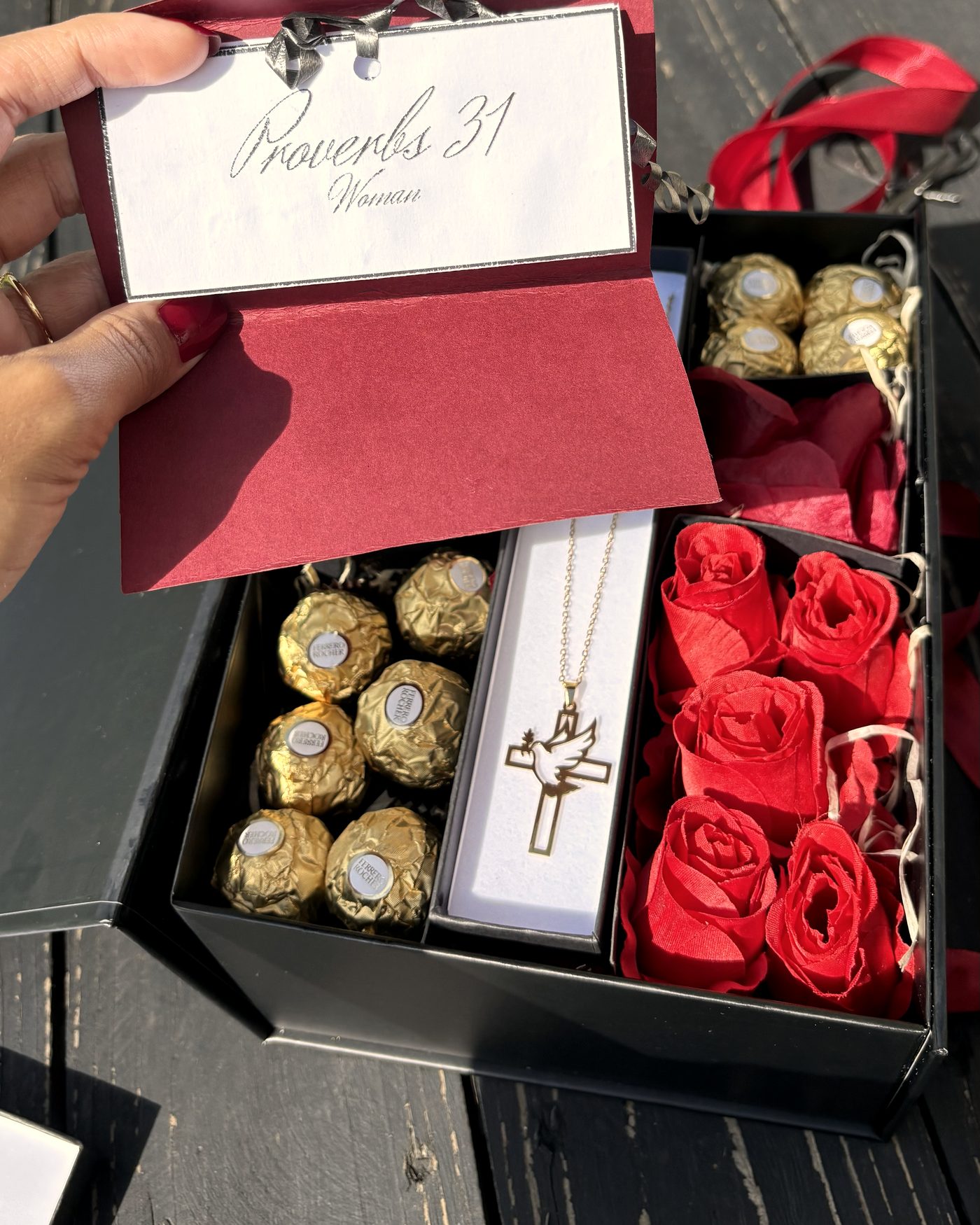 The Proverbs 31 gift box, opened — a handwritten verse card, the signature cross pendant in its case, chocolates and paper roses.